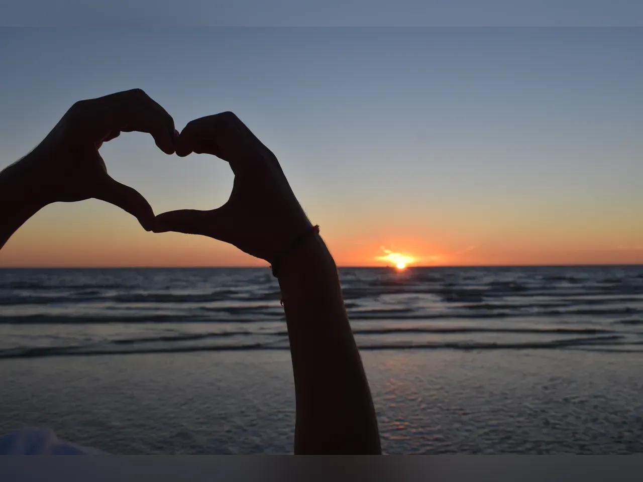 2 Hände formen ein Herz mit Meer und Sonnenuntergang im Hintergrund