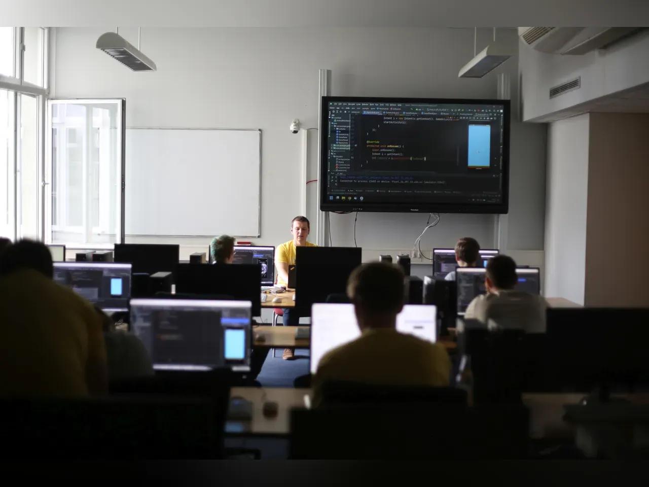 PC-Raum, an dem Menschen vor Computern sitzen, vorn wird ein Code auf einem Smartboard gezeigt