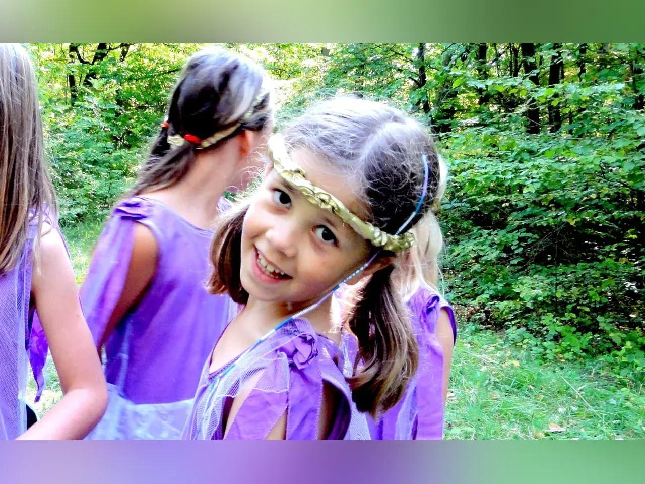 ein Mädchen im lilanen Kleid mit Kopfschmuck im Grünen mit 3 weiteren Kindern in Kostümen im Hintergrund