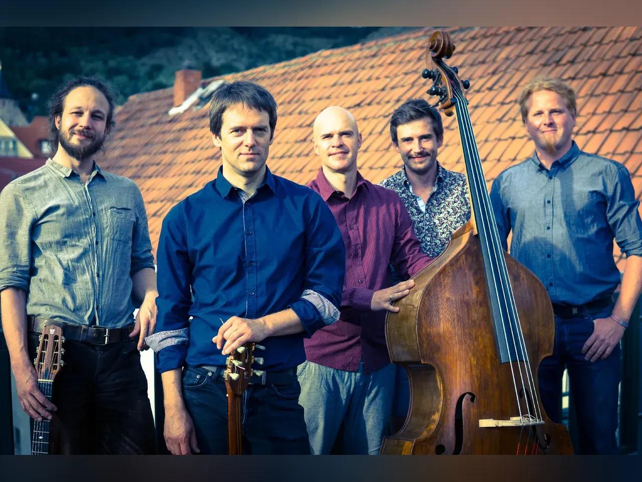 Fünf Männer der Saitensekte in Hemden und mit Kontrabass und Gitarren stehen vor einem Hausdach.
