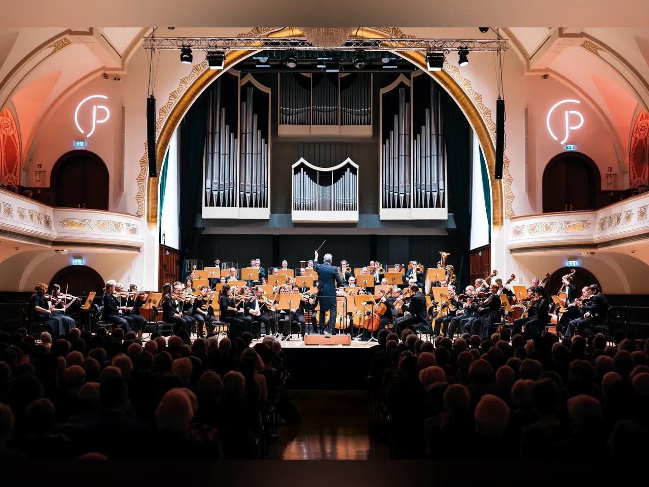 Blick auf die Bühne des Volkshauses. Der Dirigent hat den Taktstock erhoben und die Jenaer Philharmonie spielt dazu.