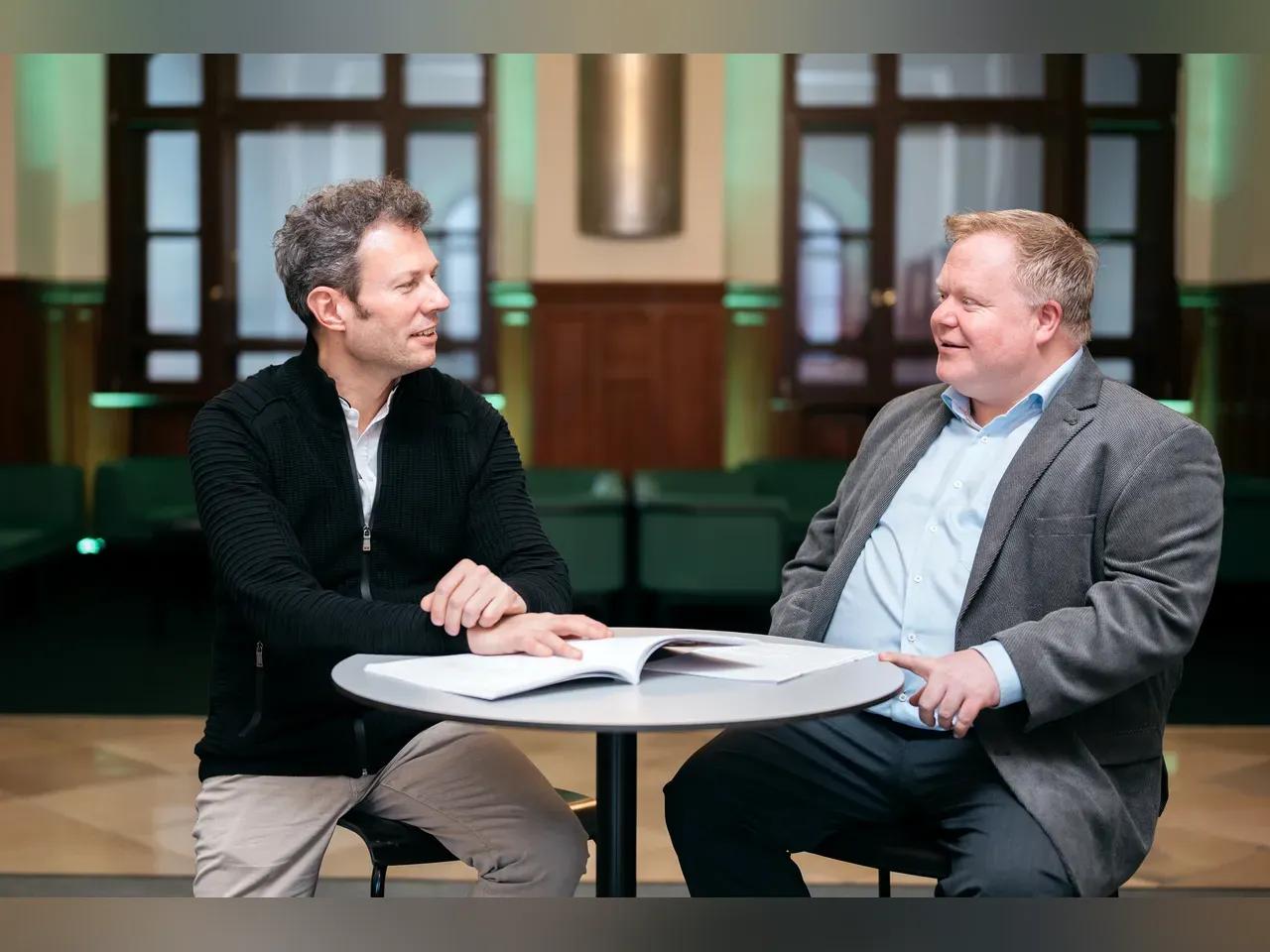 Simon Gaudenz und Alexander Richter sitzen im Foyer des Volkshauses auf Barhockern an einem Stehtisch.<br>