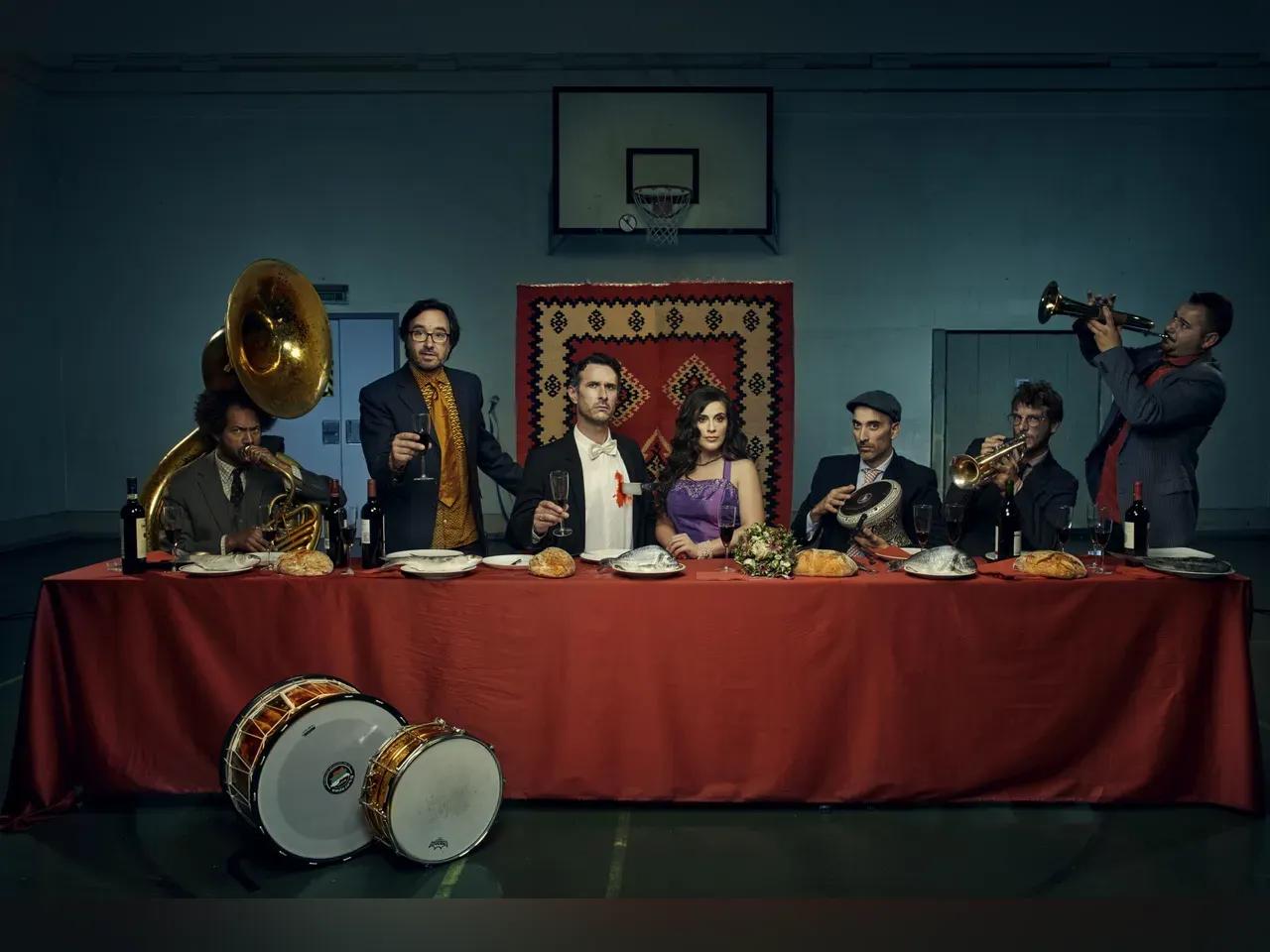 Ein Mann und eine Frau in Abendgarderobe sitzen an einer gedackten Tafel, der Mann hält sein Glas zum Anstoßen n die Höhe, auch an der Tafel sitzen Musiker mit Instrumenten.