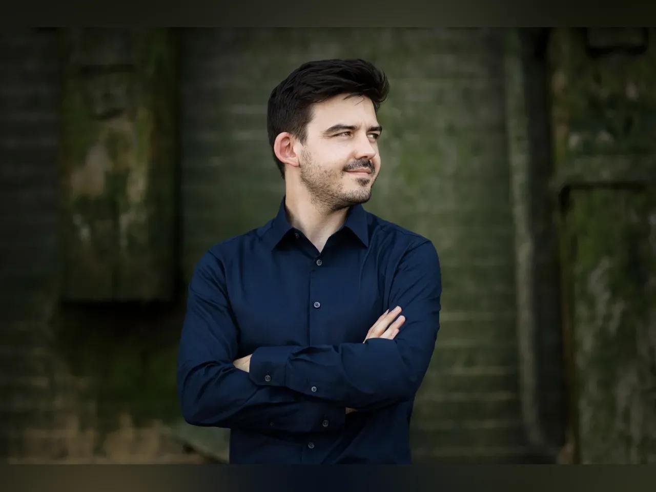 Porträtfoto des Dirigenten Harry Ogg. Er steht vor einer Wand mit verschränkten Armen und blickt nach rechts.