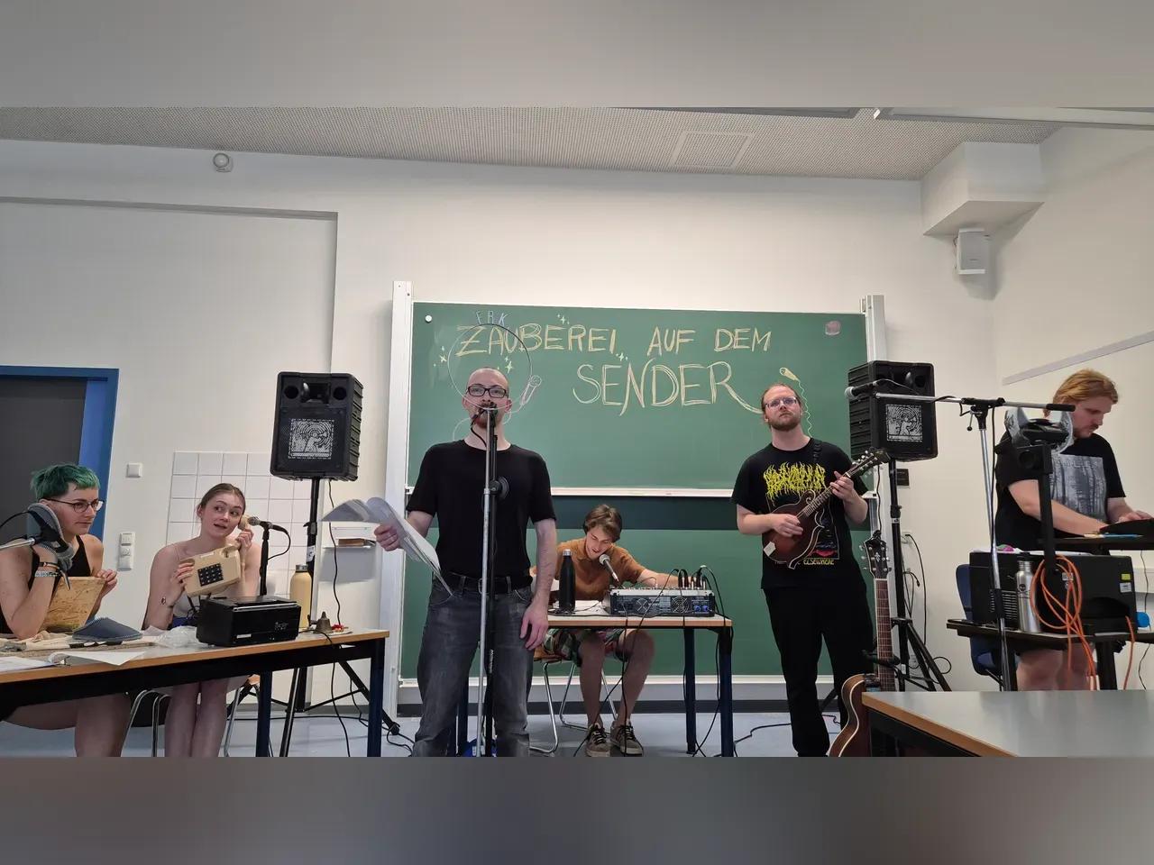 Einblick ins LIVE-Hörspiel, vor einer Tafel sitzen und stehen verschiedene Menschen mit Mikros