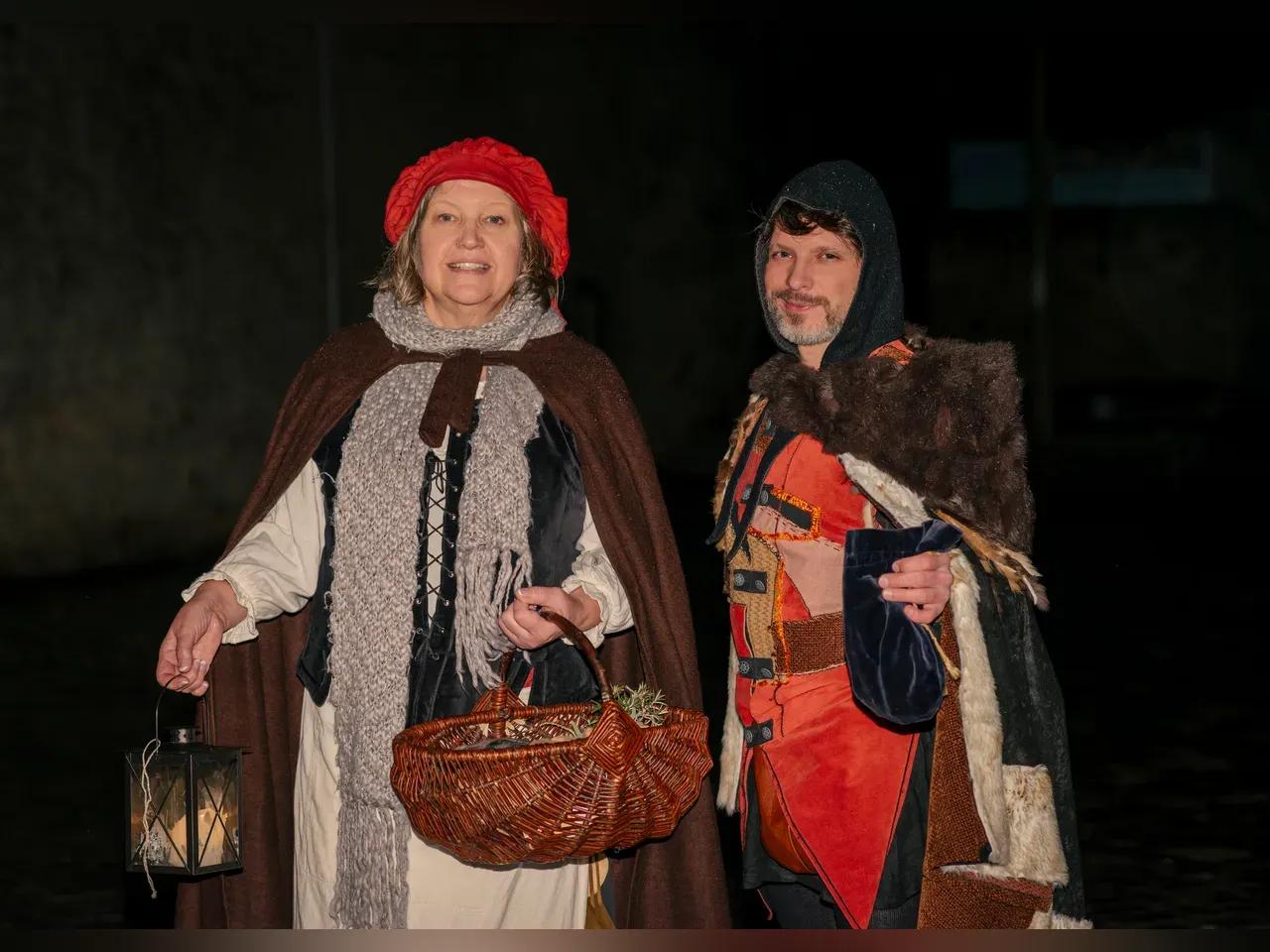 2 Personen in Kostümen bei 'Galgen,Gassen&amp;Ganoven', Frau mit rotem Hut&amp;Laterne, Mann mit Kapuzenumhang&amp;Beutel<br><br>