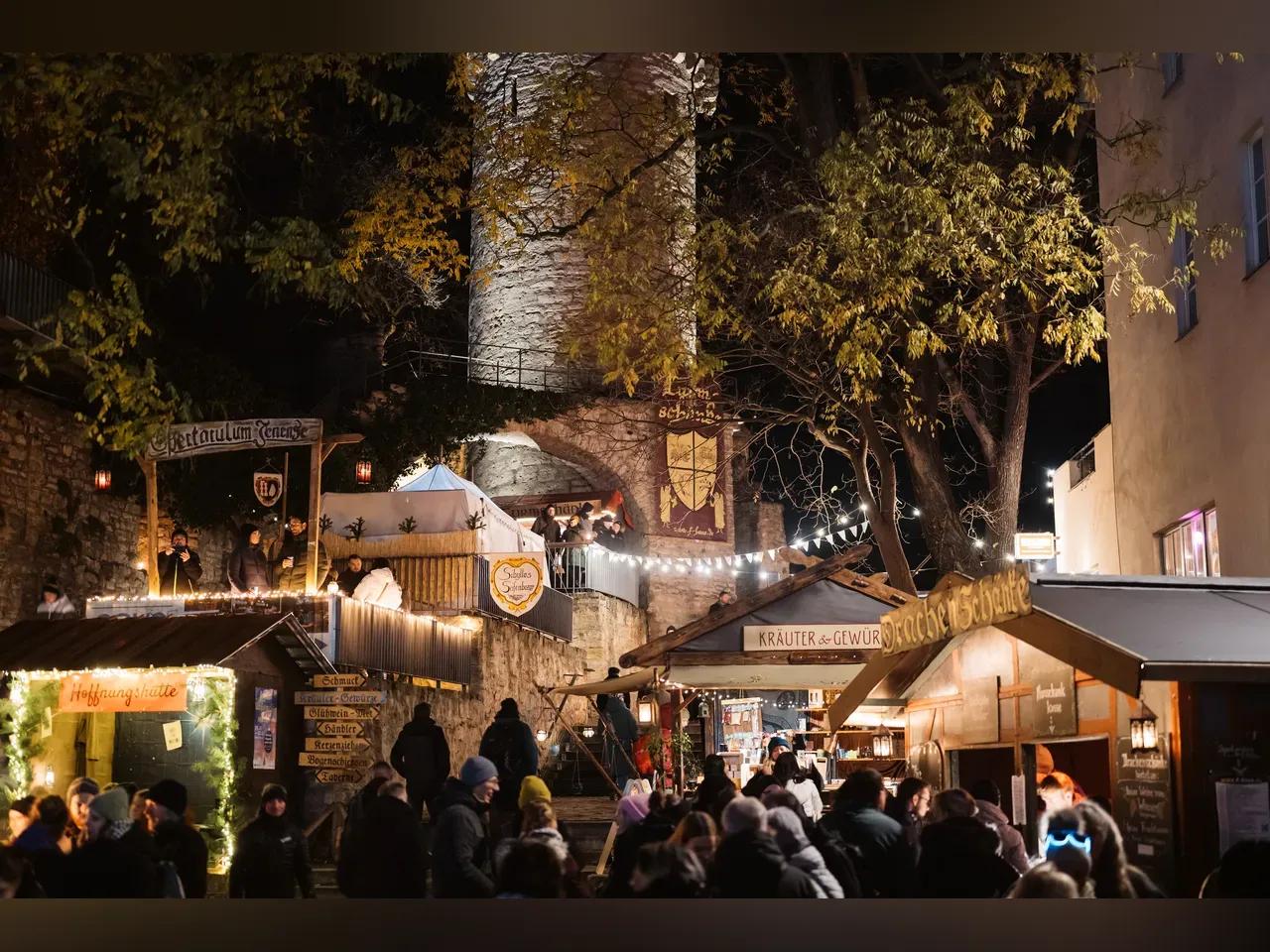 Aufnahme vom Historischen Weihnachtsmarkt mit Ständen, Feuerstellen sowie vielen Besucher:innen und einem historischem Wehturm im Hintergrund.