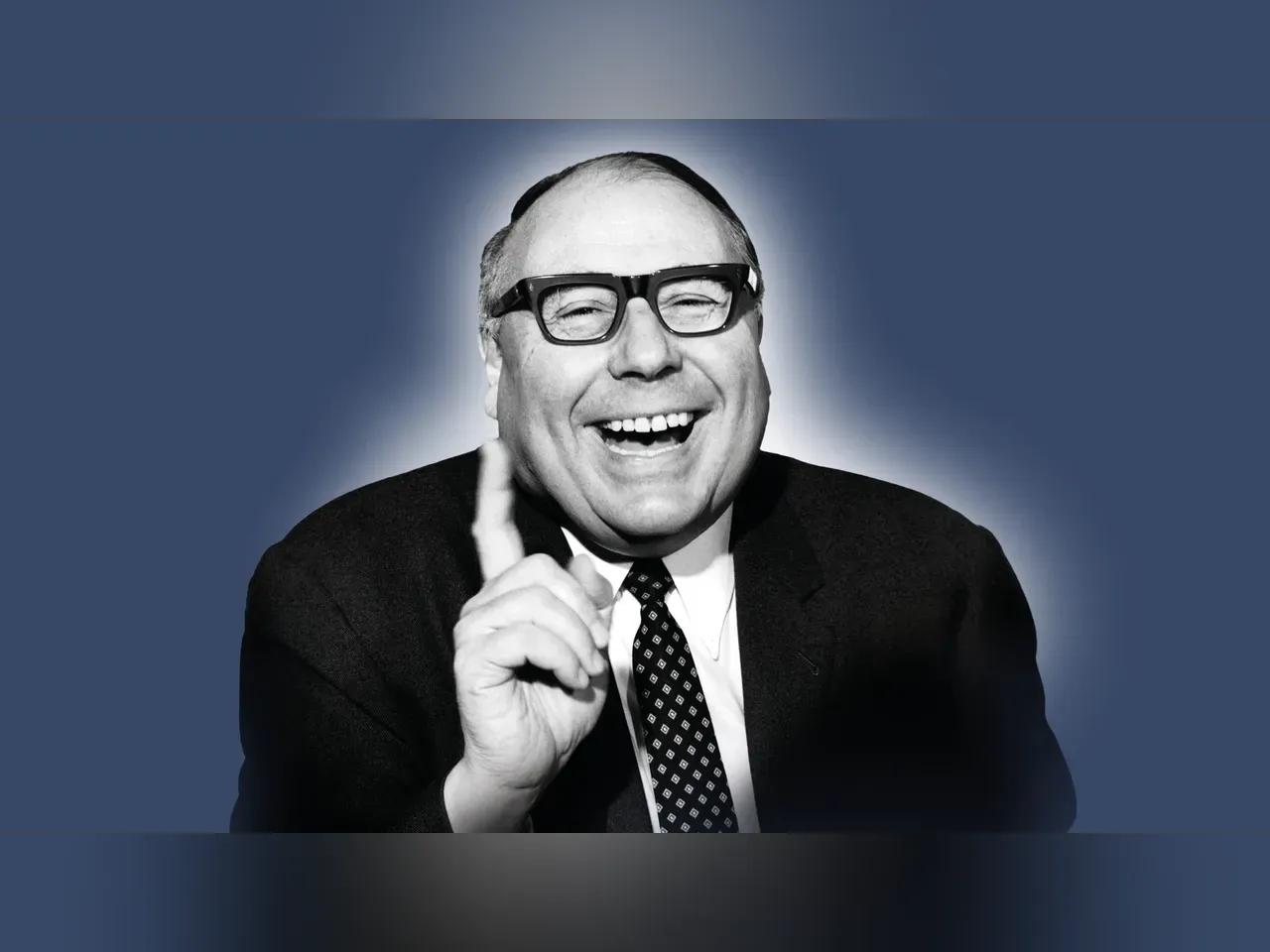 Schwarz-weiß Foto von Heinz Erhardt, lachend und mit erhobenem Zeigefinger, vor blauem Hintergrund