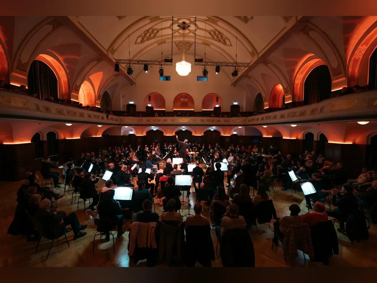 Musiker und Publikum sitzen im Kreis verteilt um ein Podest herum, auf dem Generalmusikdirektor Simon Gaudenz dirigiert.