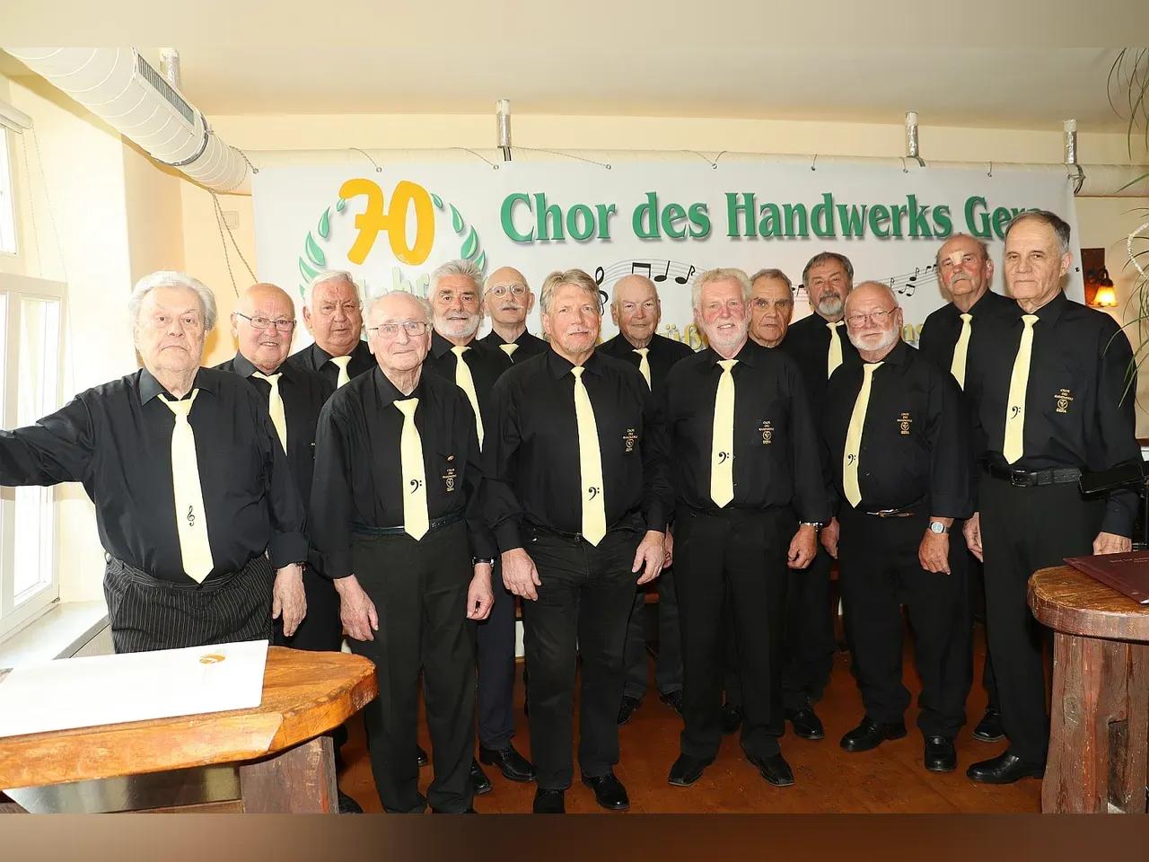 Eine Gruppe an männlich gelesenen Personen höheres Alters in schwarzen Hemden und Blusen stehen vor einen Plakat mit der Aufschrift: "70 Jahre Chor des Handwerks"