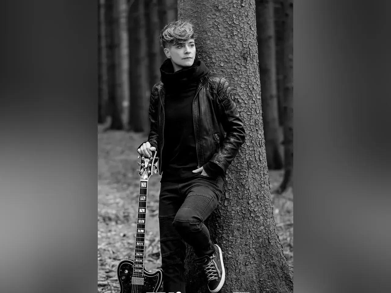 Schwarz-weiß Aufnahme von Anna Heimthal in Lederjacke, an einem Baum lehnend, die Gitarre in der Hand