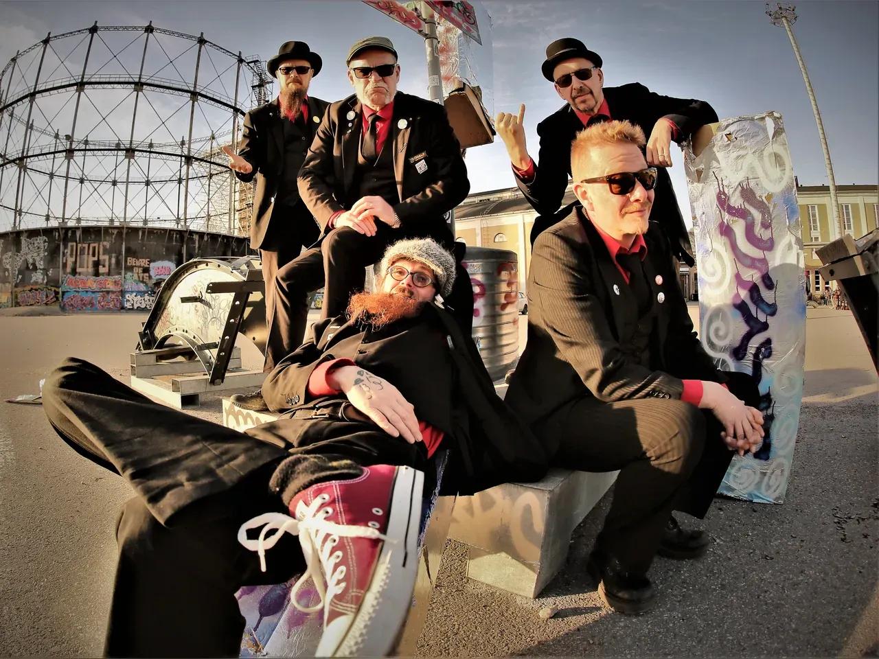 Bandmitglieder von Eläkeläiset auf einem betonierten Platz, im Hintergrund ein rundes Stahlgerüst und Graffitis