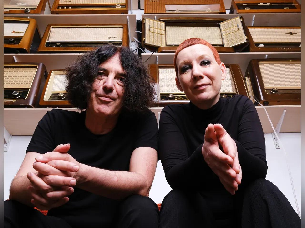 Die zwei Künstler:innen sitzen unter Regalen voller alter Radios