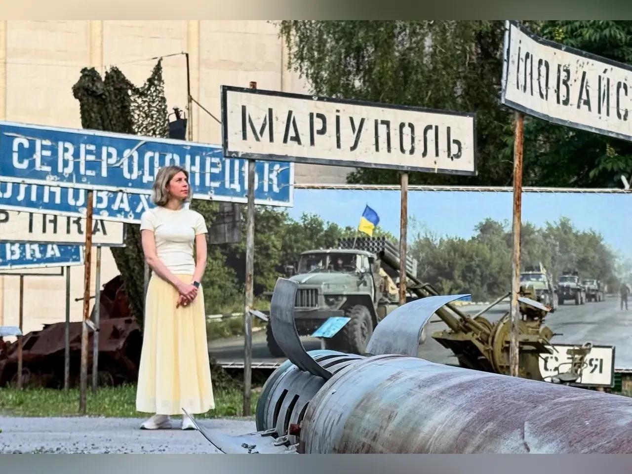 Eine Frau steht zwischen Kriegsgerät und Schildern in Ukrainischer Schrift