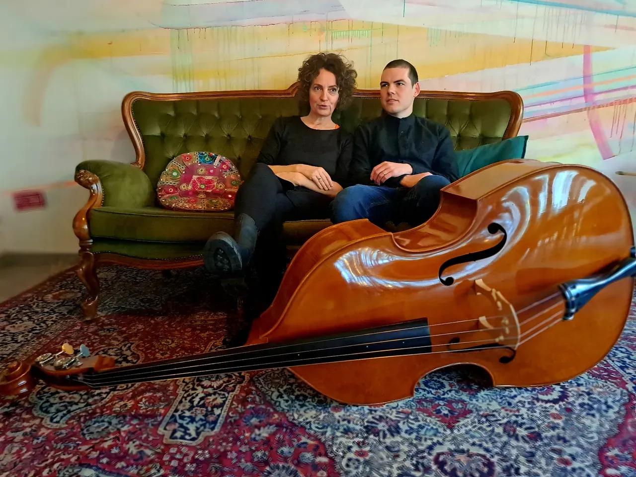Auf einem alten Barock-Sofa (mit grünem Samt) sitzen eine Frau und ein junger Mann. Vor Iihnen auf einem großen bunten, vermutlich handgeknüftem Teppich liegt ein Kontrabass. 