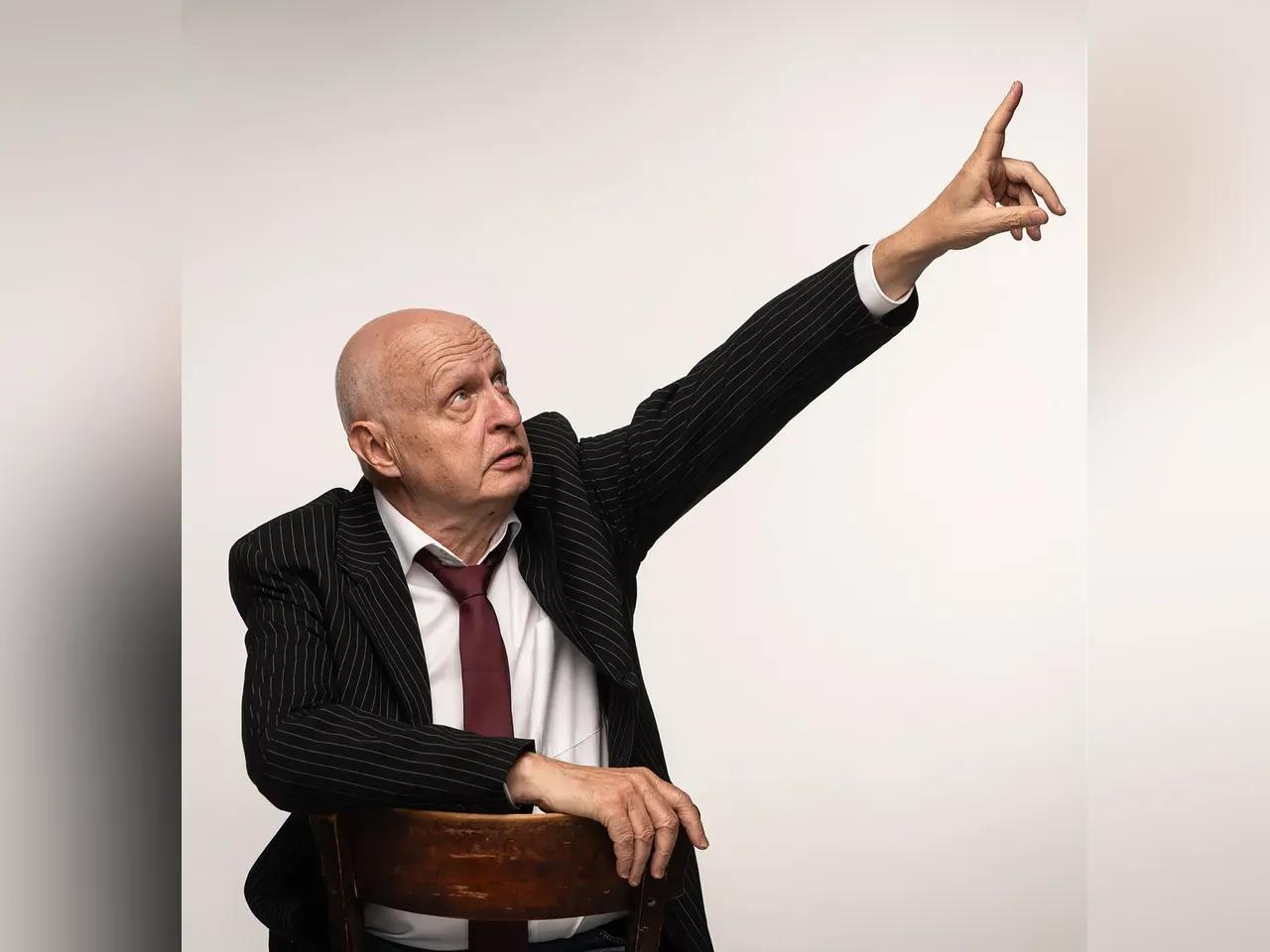 Kabarettist Heinz Klever sitzt rittlings auf einem Stuhl und zeigt mit ausgestrecktem Finger nach schräg oben