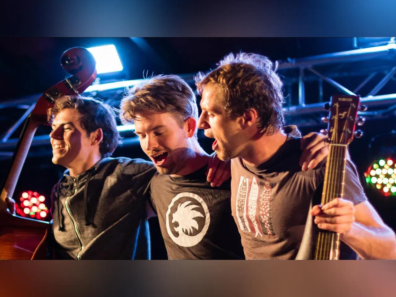 Drei Männer stehen mit Instrumenten in der Hand Arm in Arm auf der Bühne und verbeugen sich