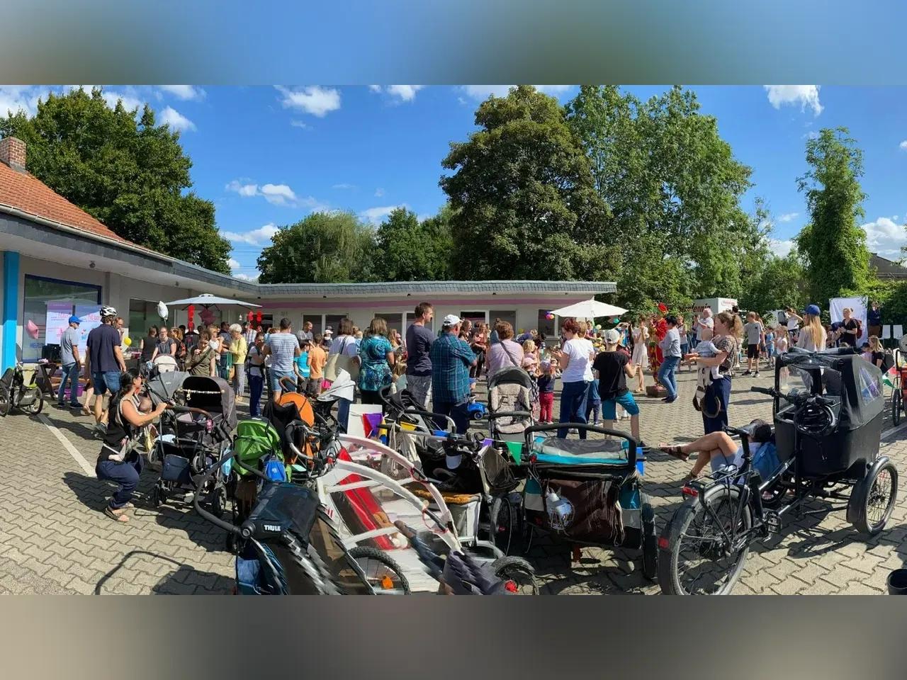 Viele Menschen auf dem Parkplatz vor einem Gebäude. Im Vordergrund sind Kinderwägen. 