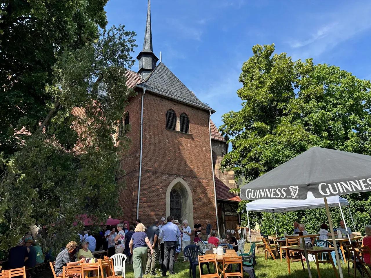 Besucher:innen, Sitzgelegenheiten und Sonnenschirm vor der Kirche Lobeda