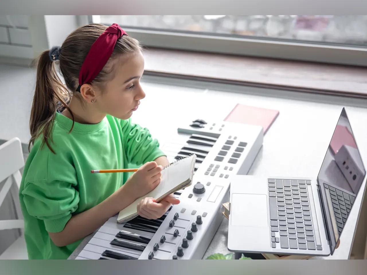 Weiblich gelesenes Kind sitzt mit Keyboard, Stift und Zettel vor Laptop