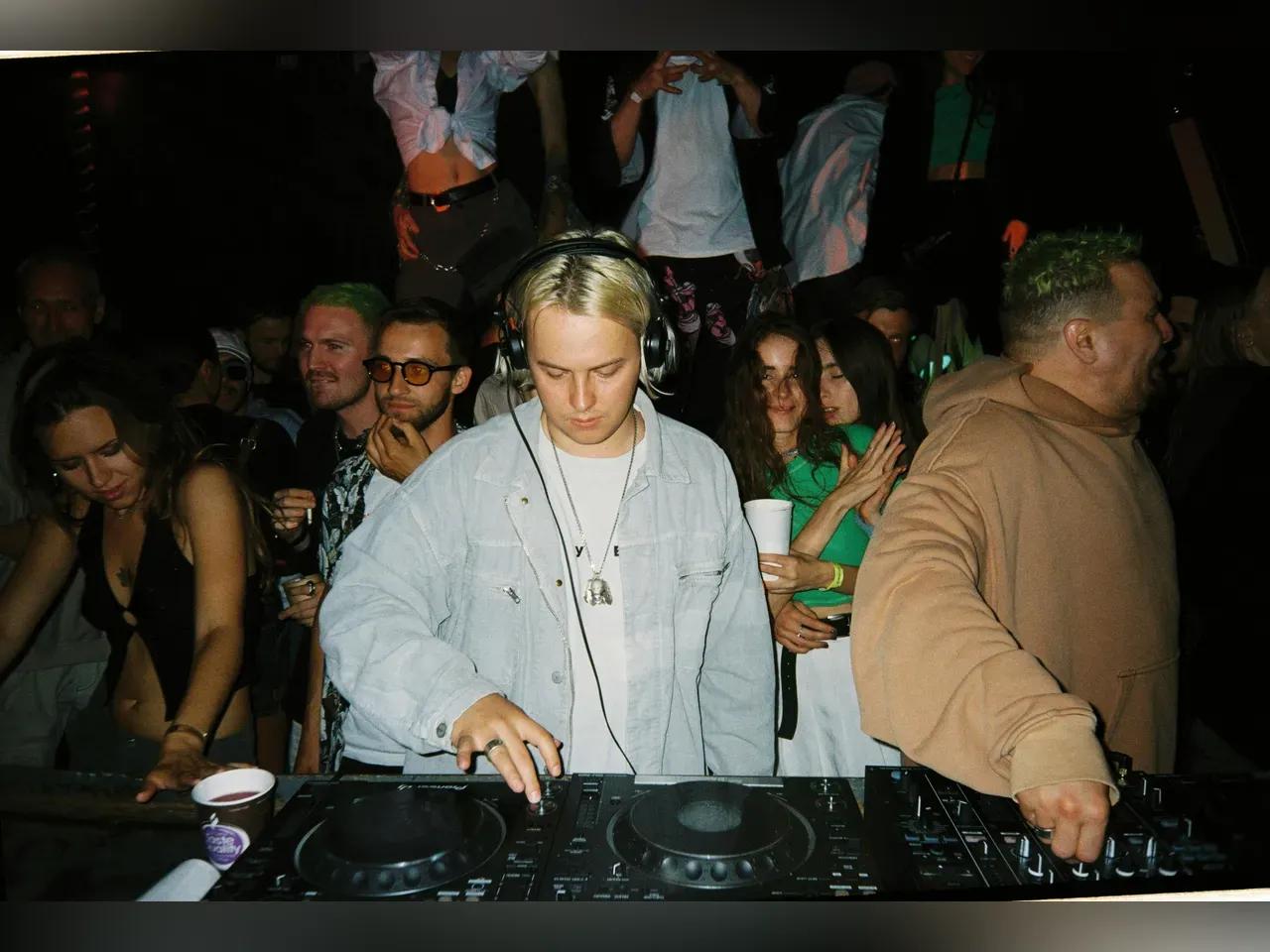 DJ steht am Pult und mixt, im Hintergrund tanzen Menschen