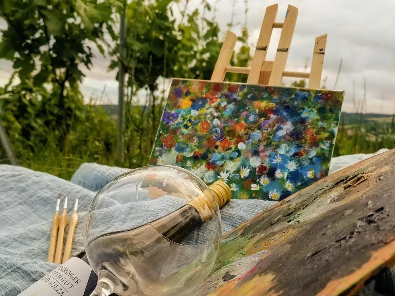 In einem Weinberg steht eine Staffelei mit einem Bild, davor liegen auf einer Decke Pinsel, eine flasche Wein, ein Weinglas und eine Farbpalette