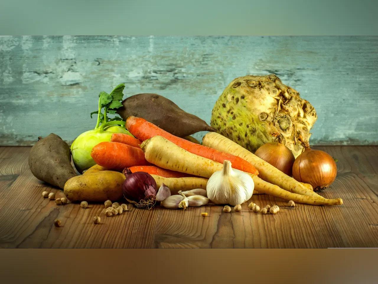 Verschiedene Gemüsesorten wir Rettich, Pastinaken, Möhren, Kohlrabi, Zwiebeln usw. liegen auf gehäuft auf einem Holztisch. Der Hintergrund ist eine bläuliche Steinwand.