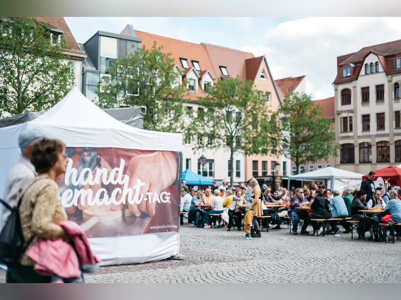 Zwei Menschen laufen an einem Pavillon vorbei, an dem ein Banner gehängt wurde, an dem darauf steht "handgemacht-Tage", im Hintergrund sitzen mehrere Menschen auf Bierbänken.
