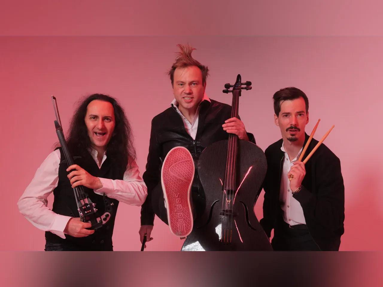 Die drei Bandmitglieder von Stilbruch mit Geige, Cello und Drumsticks vor rosa Hintergrund