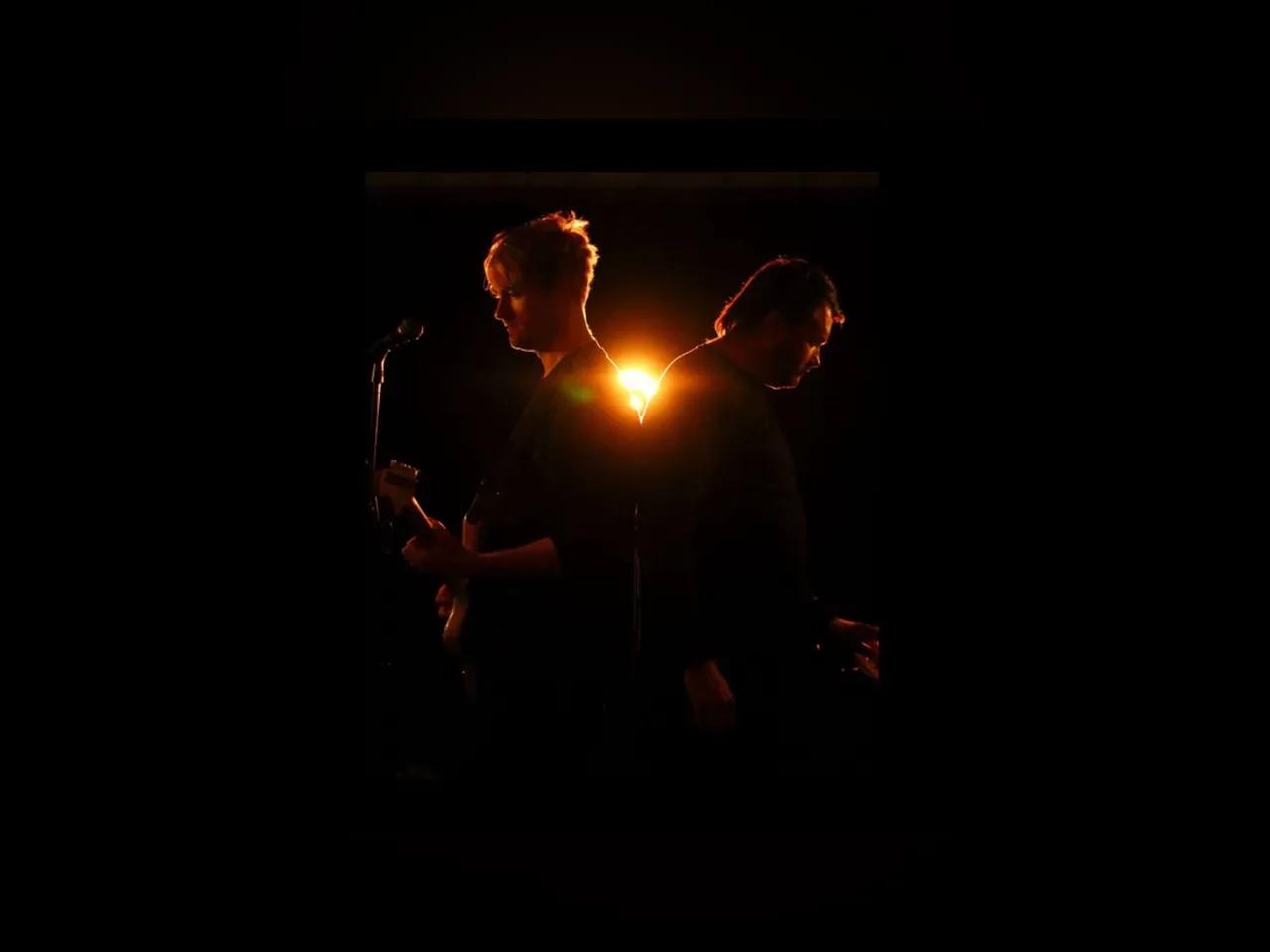 Silhouetten der zwei Musiker vor schwarzem Hintergrund mit einem orangenen Licht